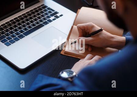 Businessman writing note with pen inspired ideas list on notepad while working on laptop computer in office, dark tone, close up. Man attending online Stock Photo