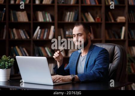 A young businessman with glasses is working on a laptop in a luxurious office in the library. A bearded man in a blue suit at a Desk is typing on a Stock Photo