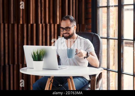 Young successful bearded businessman freelancer in office coworking. Happy man programmer developer in a white t shirt smiling and drinking coffee at Stock Photo