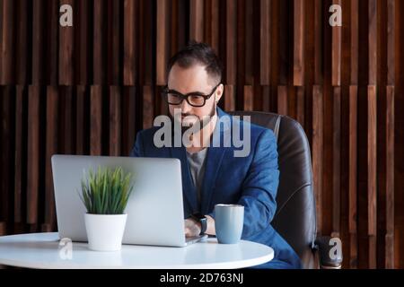 Young successful bearded businessman freelancer in office coworking. Happy man programmer developer in a white t shirt smiling and drinking coffee at Stock Photo