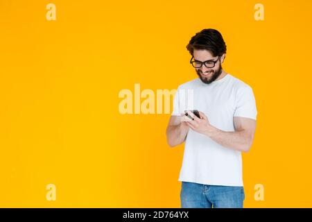 Close-up of his cute attractive cheerful bearded boyfriend in white shirt looking at phone screen isolated on bright vivid bright yellow background Stock Photo