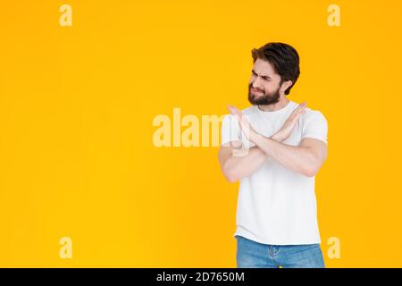 A serious young bearded man in his 30s in a white t-shirt poses standing up showing a stop gesture with his arms crossed looking at the camera Stock Photo