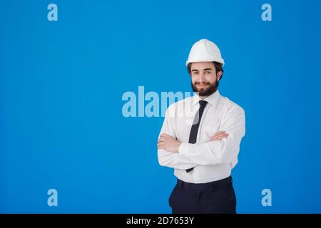 Male Engineer architect in a white shirt and construction helmet on a blue background , Studio portrait. The construction worker crosses his arms and Stock Photo