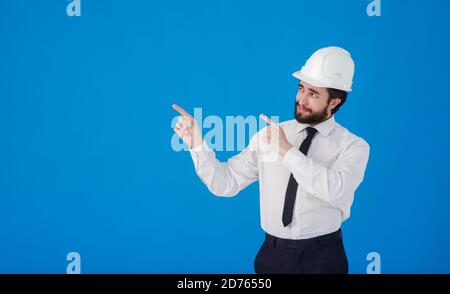 Handsome man industrial engineer wearing a white hard helmet and pointing finger towards something blank copy space for text isolated on blue Stock Photo
