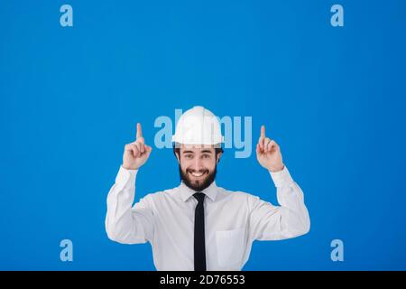 Handsome man industrial engineer wearing a white hard helmet and pointing finger towards something blank copy space for text isolated on blue Stock Photo