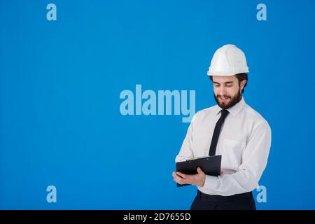 A young bearded male construction worker in a white shirt and construction helmet on a blue background. industrial engineer-Architect hold clipboard Stock Photo