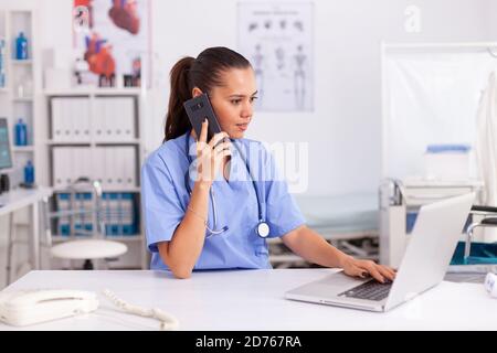 Medical practitioner using telephone and laptop in hospital office wearing blue uniform. Health care physician sitting at desk using computer in modern clinic looking at monitor. Stock Photo