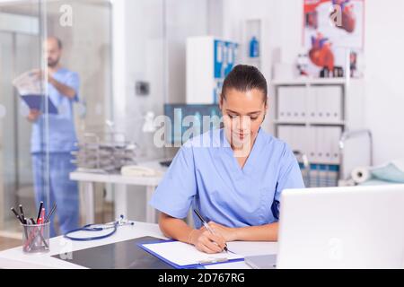 Healhcare practitioner checking patient insurance on laptop and taking notes. Health care physician using computer in modern clinic looking at monitor, medicine, profession, scrubs. Stock Photo