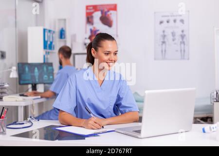 Medical practitioner sitting at desk in hospital office using laptop with doctor in the background. Health care physician using computer in modern clinic looking at monitor, medicine, profession. Stock Photo