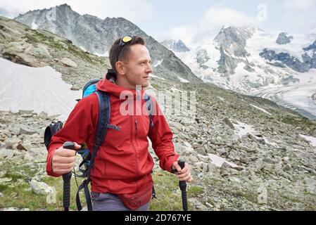 Portrait of a man alpinist wearing a red jacket and a backpack, having a walk with hiking poles in the mountains in Swiss Alps, amazing landscape around him