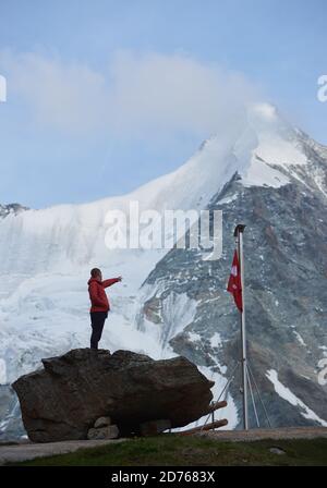 Male tourist looking at beautiful mountains scenery and pointing with finger. Man reaching peak, standing on stones near the flagpole with Swiss flag. Nature with amazing views. Sport tourism in Alps. Stock Photo