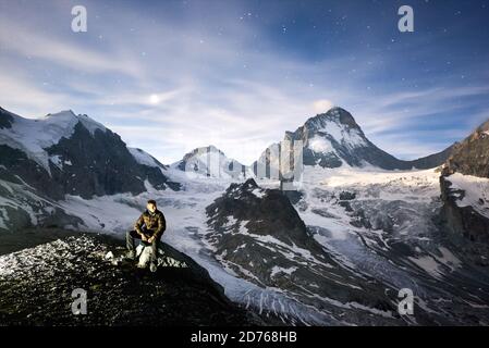 Amazing scenery in evening, man sitting on stone, beautiful mountains with white snow on background. Gorgeous mountain ridge with high rocky peaks, milky way with shining stars in sky, wonderland. Stock Photo