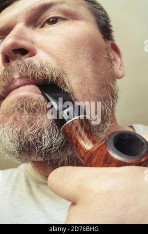 Unusual portrait of a bearded man with a smoking pipe in his mouth. A grown man with gray stubble and a heather-root smoking pipe. Grimaces in front o Stock Photo