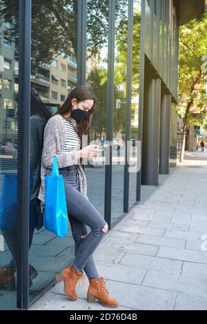 Young woman wearing face mask using her smartphone leaning on a shop window. New normal concept. Stock Photo