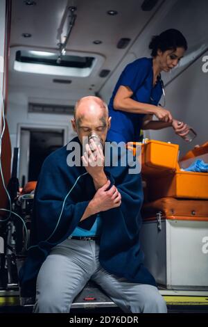 Injured man in a blanket breathing through an oxygen mask in an ambulance car, nurse searching for something in her medical kit in the background. Stock Photo