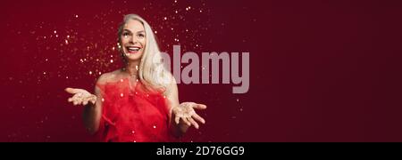 Happy holidays with excited senior woman throwing glitters in studio. Caucasian senior woman playing with golden star glitters over red background. Stock Photo