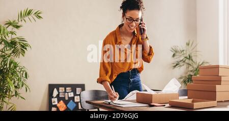 Drop shipping business owner talking on mobile phone and taking order. Female entrepreneur working at home office confirming the order on phone. Stock Photo