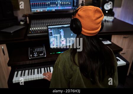 Young woman, female artist looking focused while playing keyboard synthesizer, creating music, sitting in recording studio Stock Photo