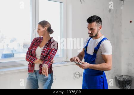 A builder in a blue overalls shows the customer the renovation of an apartment. He writes notes with a pen Stock Photo