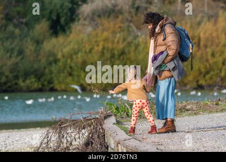 Adult and child standing wearing coats on a cold Autumn morning, feeding birds and ducks on a lake in the UK. Stock Photo