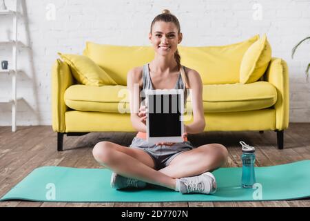 Smiling sportswoman showing digital tablet with blank screen near sports bottle on fitness mat