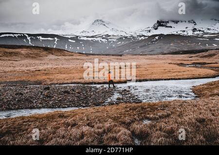 Adventurous man walking and hopping on rocks across shallow area of a river, Ice melts on a river in the mountains, with ice mountain in the backgroun Stock Photo