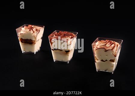 Three small glassed with dessert in row decorated with whipped cream and cocoa, isolated on black. Closeup of tiramisu cake with three layers of chocolate biscuit and natural coffee syrup mousse. Stock Photo