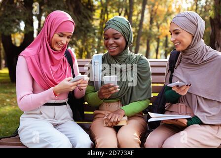Female Muslim Students Using Smartphone Phones Sitting In Park Outside Stock Photo