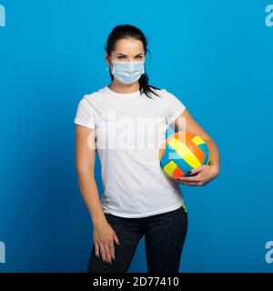 Happy sporty woman with face mask from coronavirus holding ball. Isolated on blue Stock Photo