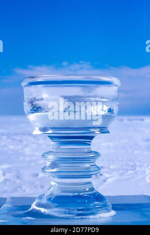 Ice bowl cut out of ice against the background of snow and blue sky. A beautiful ice cup stands on an ice pedestal. Cracks and scratches on the ice. Stock Photo