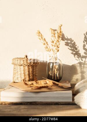 Cozy autumn home still life of dry flowers in vase, straw box, knitted blanket and magazines on rustic wooden table and shadows on the wall from the b Stock Photo