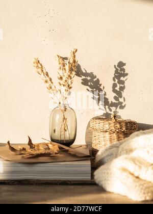 Cozy autumn home still life of dry flowers in vase, straw box, knitted blanket and magazines on rustic wooden table and shadows on the wall from the b Stock Photo