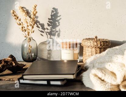 Cozy autumn home still life of dry flowers in vase, straw box, glass candle, knitted blanket and black notepads on rustic wooden table and shadows on Stock Photo