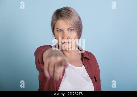 Serious caucasian senior woman is pointing at camera with forefinger posing on a blue studio wall in casual clothes Stock Photo