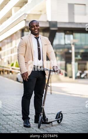 Happy african-american businessman is standing with scooter in front of company building. Stock Photo