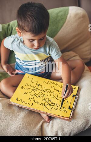 Cute caucasian boy is drawing something on a yellow paper with a marker while sitting at home in bed Stock Photo