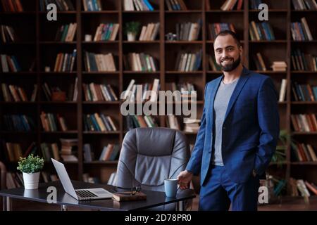 Young bearded businessman in a blue suit in the office library. A man stands at the desktop with a laptop Stock Photo