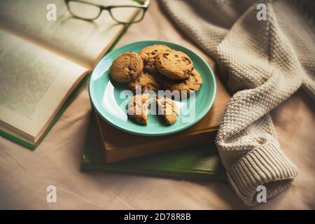 A blue plate of homemade chocolate cookies are on top of a stack of antique books, next to which is an open book, glasses, and the beige sleeve of a w Stock Photo