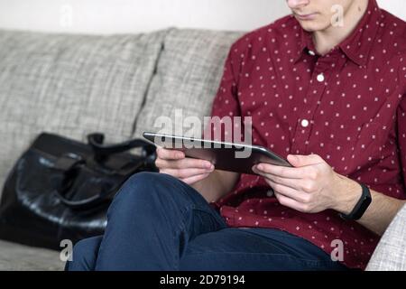 The young man sat down on the sofa in the office with his legs crossed. Looks at the information on the tablet, put a briefcase next to it Stock Photo