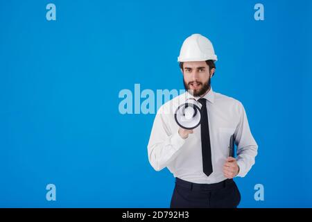 A young bearded male construction worker in a white shirt and construction helmet on a blue background. An industrial engineer-Architect shouts Stock Photo