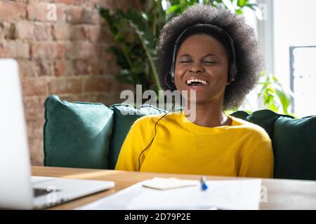 Call support can't stop laughing. Smiling African American millennial woman wear headphones while talking to a client in video chat, teach online, wat Stock Photo
