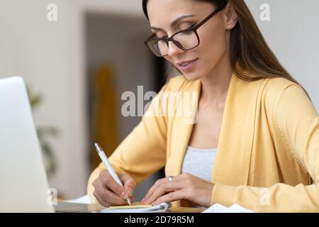 Focused business woman in eyeglasses wear yellow cardigan makes a note in notebook. Office female manager remotely works from home during the second w Stock Photo