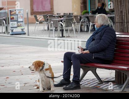 An elderly man wearing a face mask sitting on a bench in the square in the town enjoying an afternoon with his dog and complying with coronavirus. Stock Photo