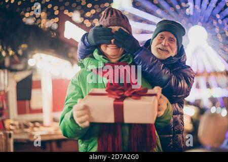 Photo of two people retired lovers couple palms cover eyes grandpa prepare giftbox grandma hold white box red bow wear coat red scarf headwear x-mas Stock Photo