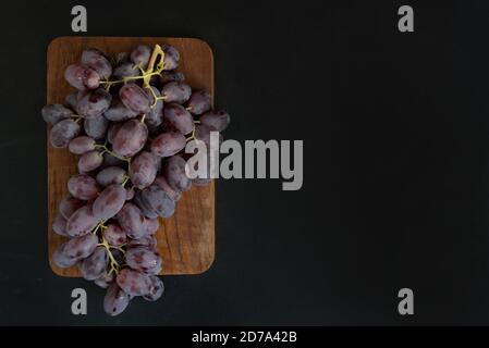 A bunch of purple grapes on a serving wooden board against a dark background with space for text. Poster. Stock Photo