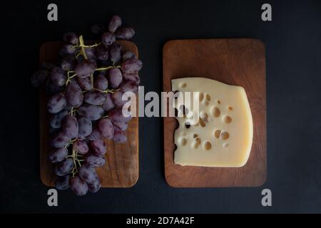 Maasdam cheese and a bunch of purple grapes on serving wooden boards against a dark background. Poster. Stock Photo