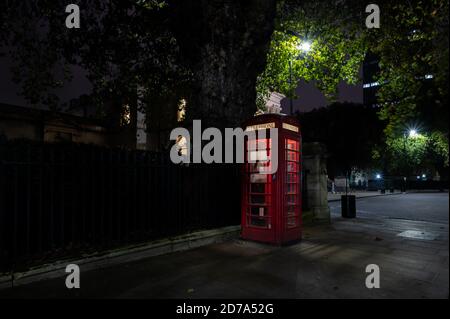 London, UK - 18 Oct 2020: British red telephone box in London in front of trees and black railings Stock Photo