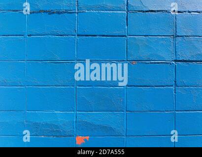 Surface of an old concrete block wall painted bright blue with areas of peeling paint showing orange underneath. Stock Photo