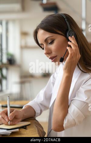 Focused woman consultant in shirt wear headphones communicating with clients, talking on video conference, makes a note in notebook, remote work from Stock Photo