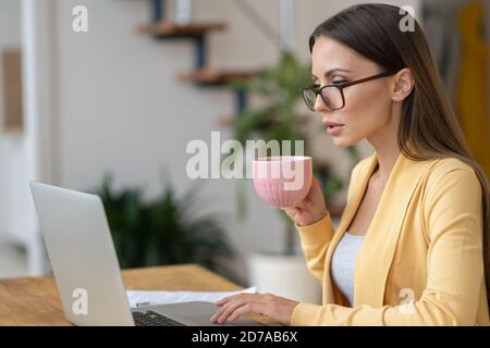 Focused millennial woman in glasses sitting on table, holding mug of tea, remote work on laptop, analyzing project statistics information, replies to Stock Photo